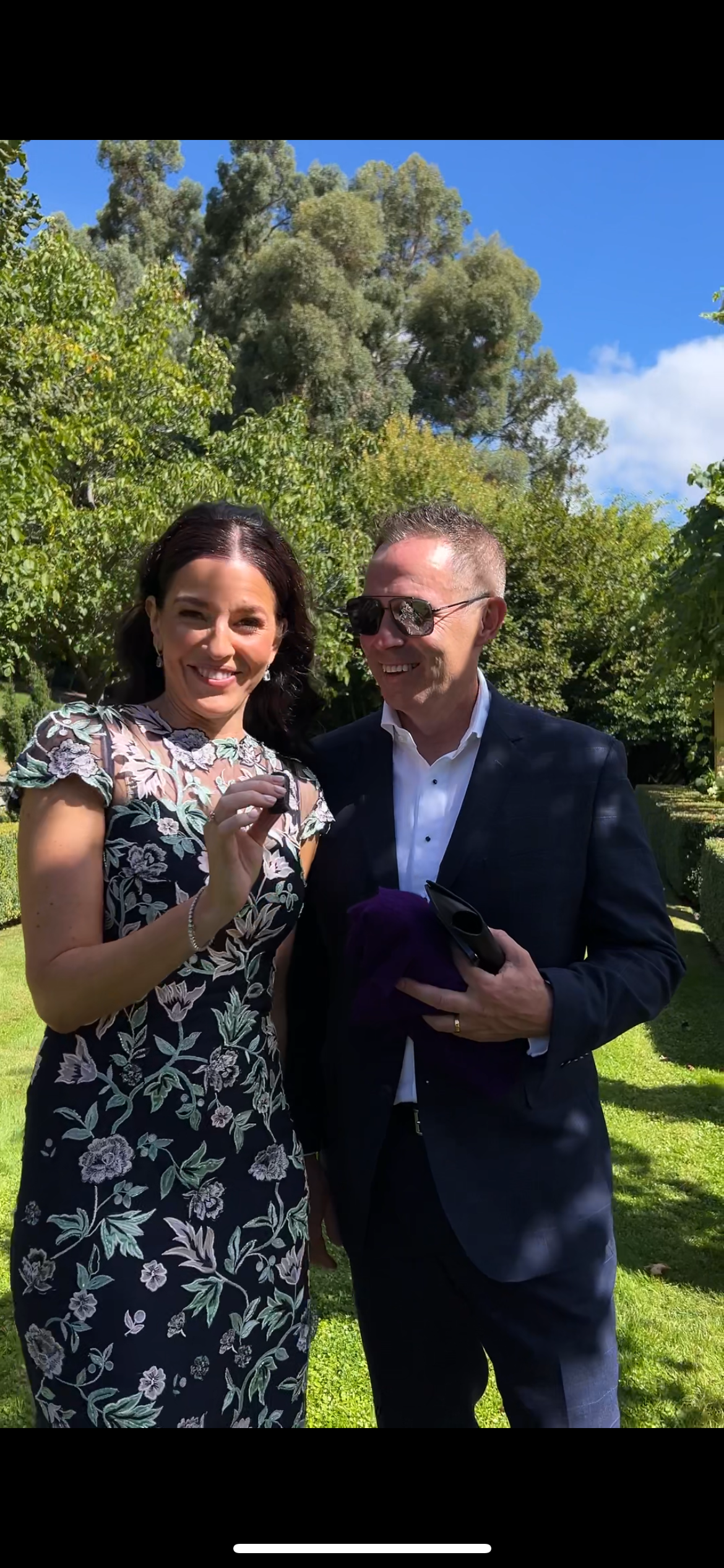 A couple in formal attire poses together outdoors on a sunny day with trees and greenery in the background.