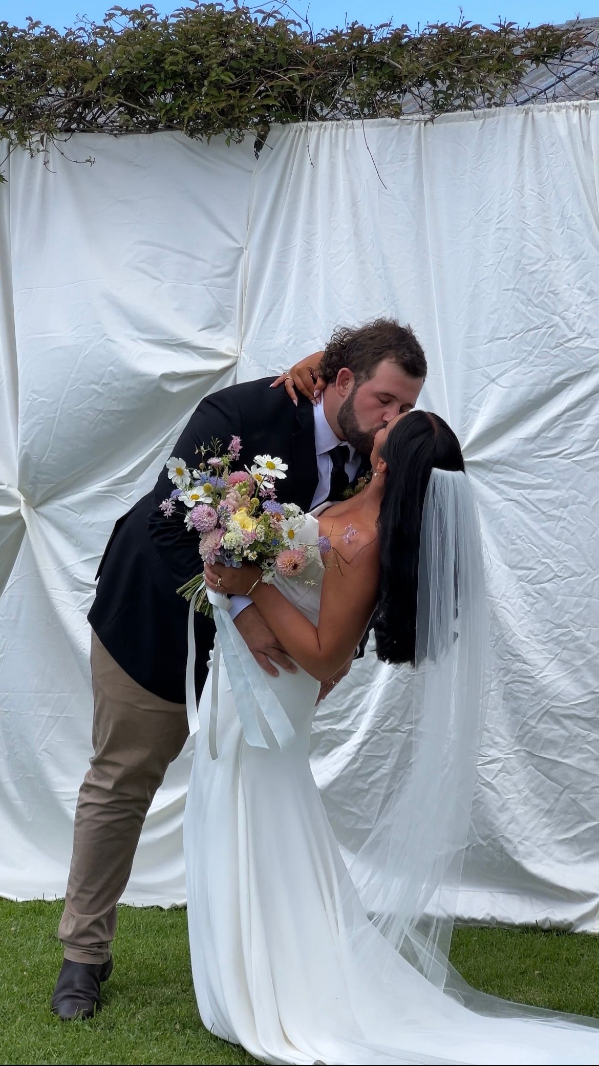 Three sequential photos show a couple in wedding attire sharing a romantic kiss behind a white curtain outdoors.
