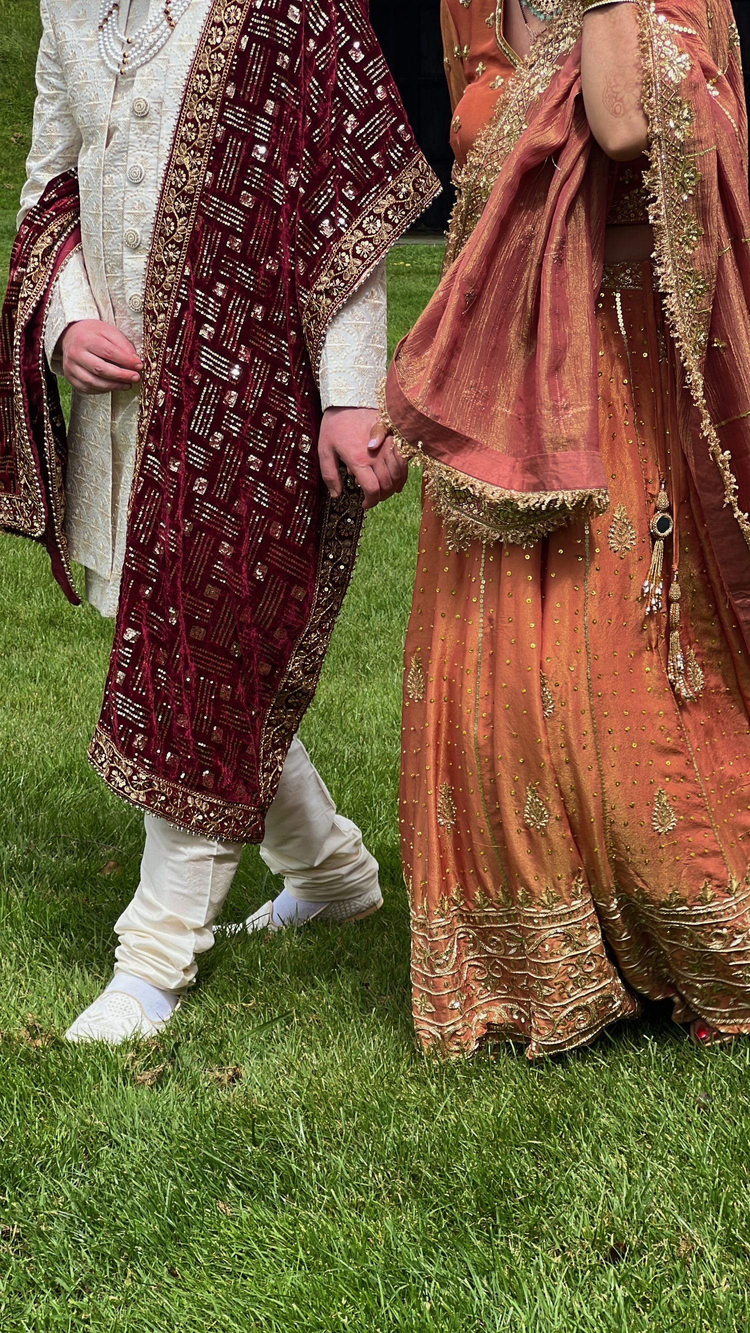 Traditional Indian wedding attire with burgundy shawl and coral lehenga shown from waist down on grass.