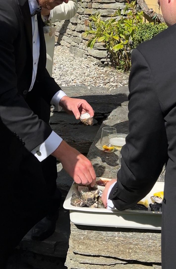 Person in formal attire preparing food on a white plate outdoors against a stone wall backdrop.