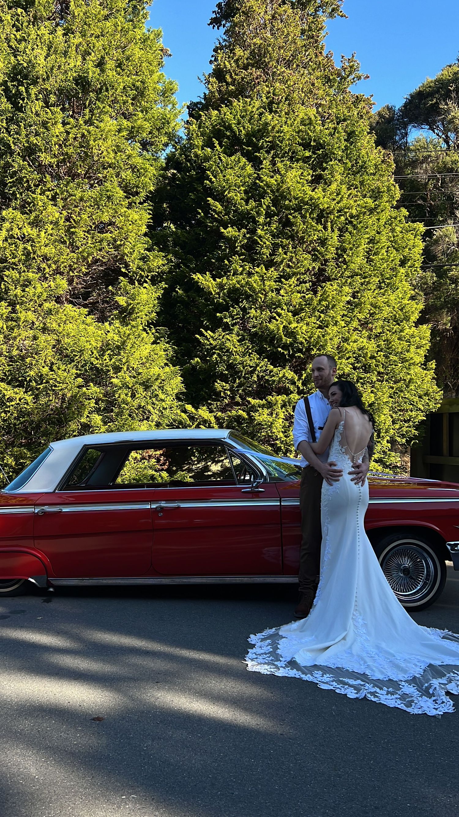 A couple in wedding attire poses next to a classic red vintage car on a sunny day.