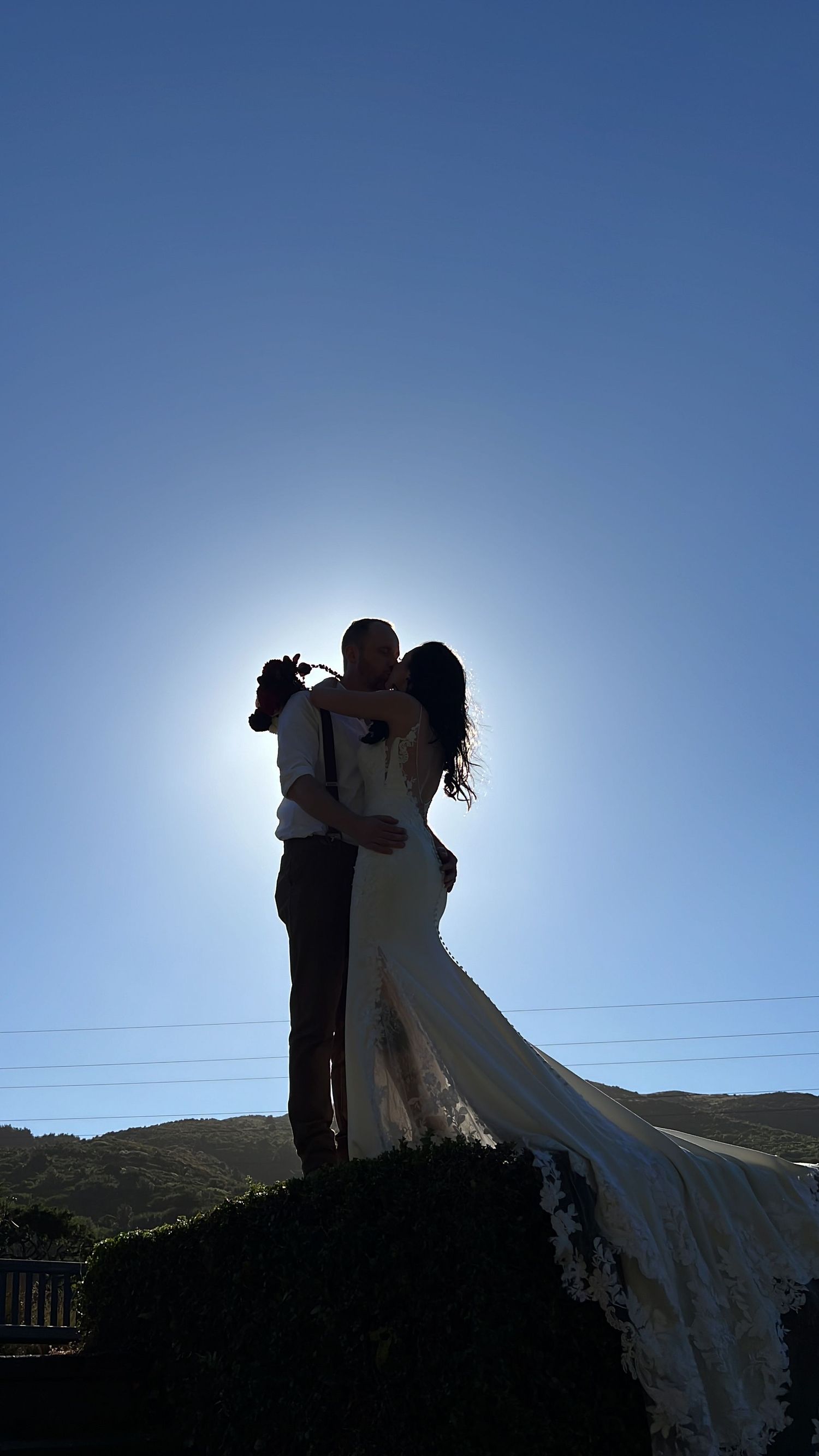 Silhouetted couple embracing against a bright blue sky on a hilltop.