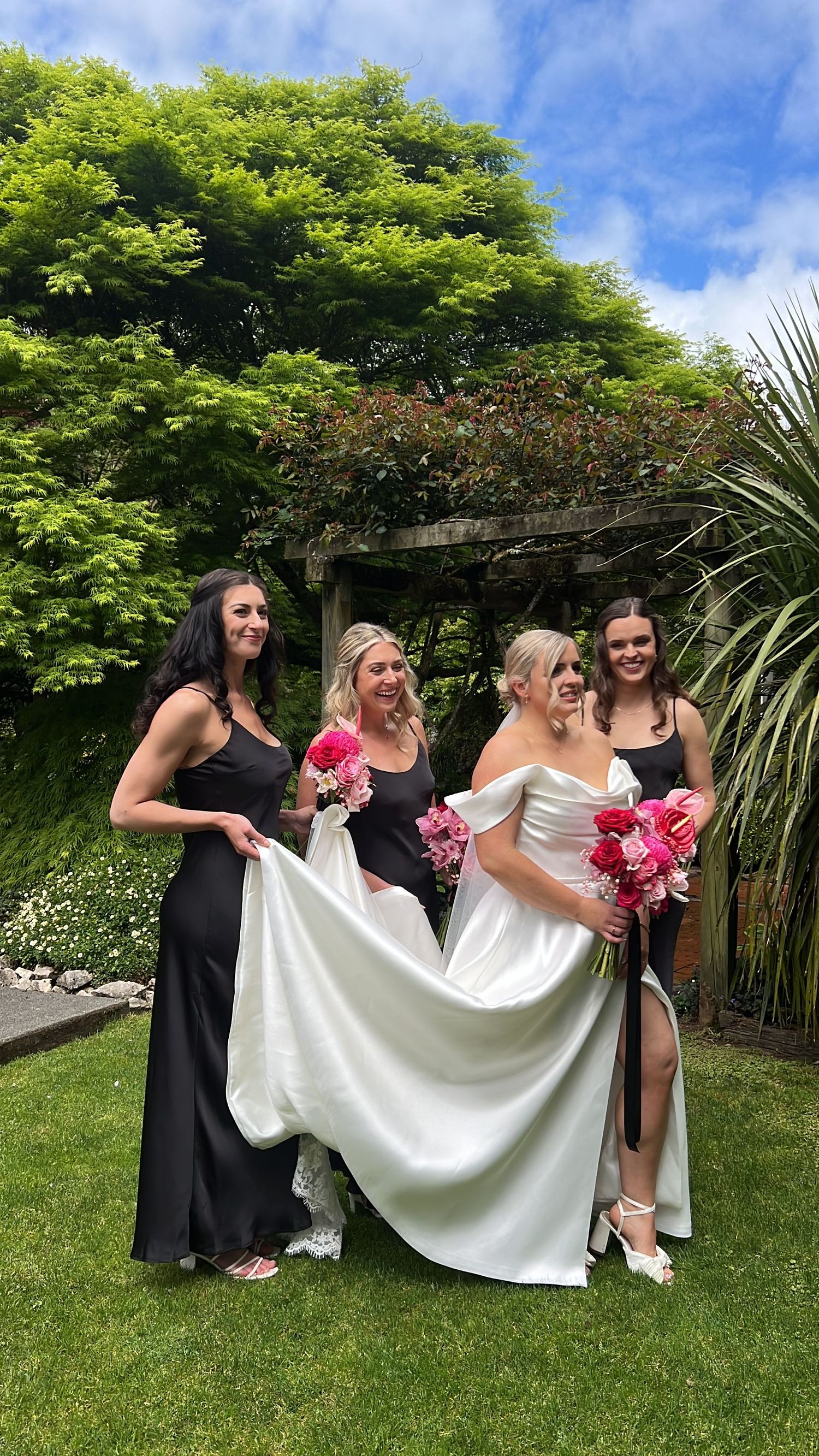 Wedding party arranges bride's dress on sunny lawn with garden backdrop.