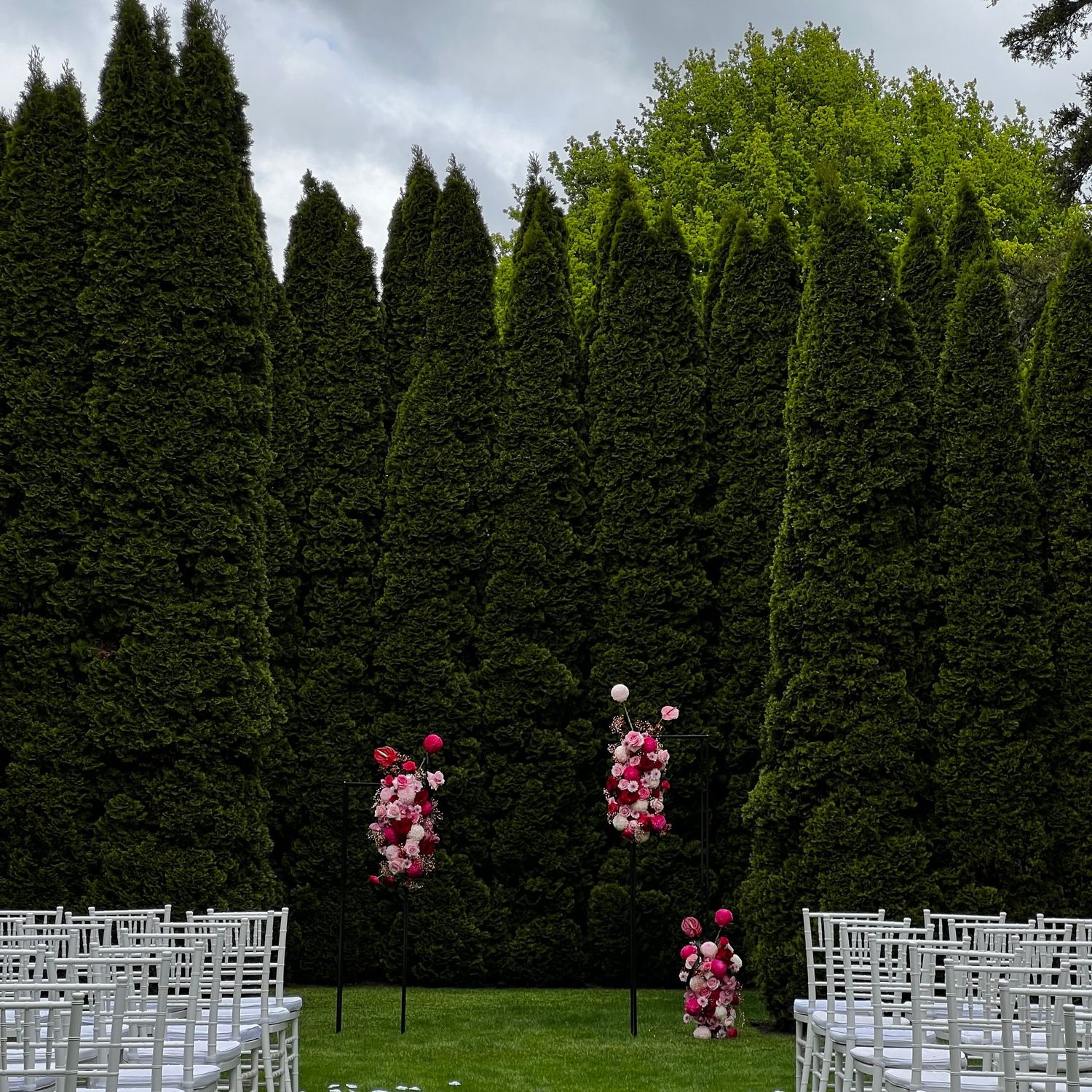 White chairs set up for an outdoor wedding ceremony with pink floral decorations and cypress trees.