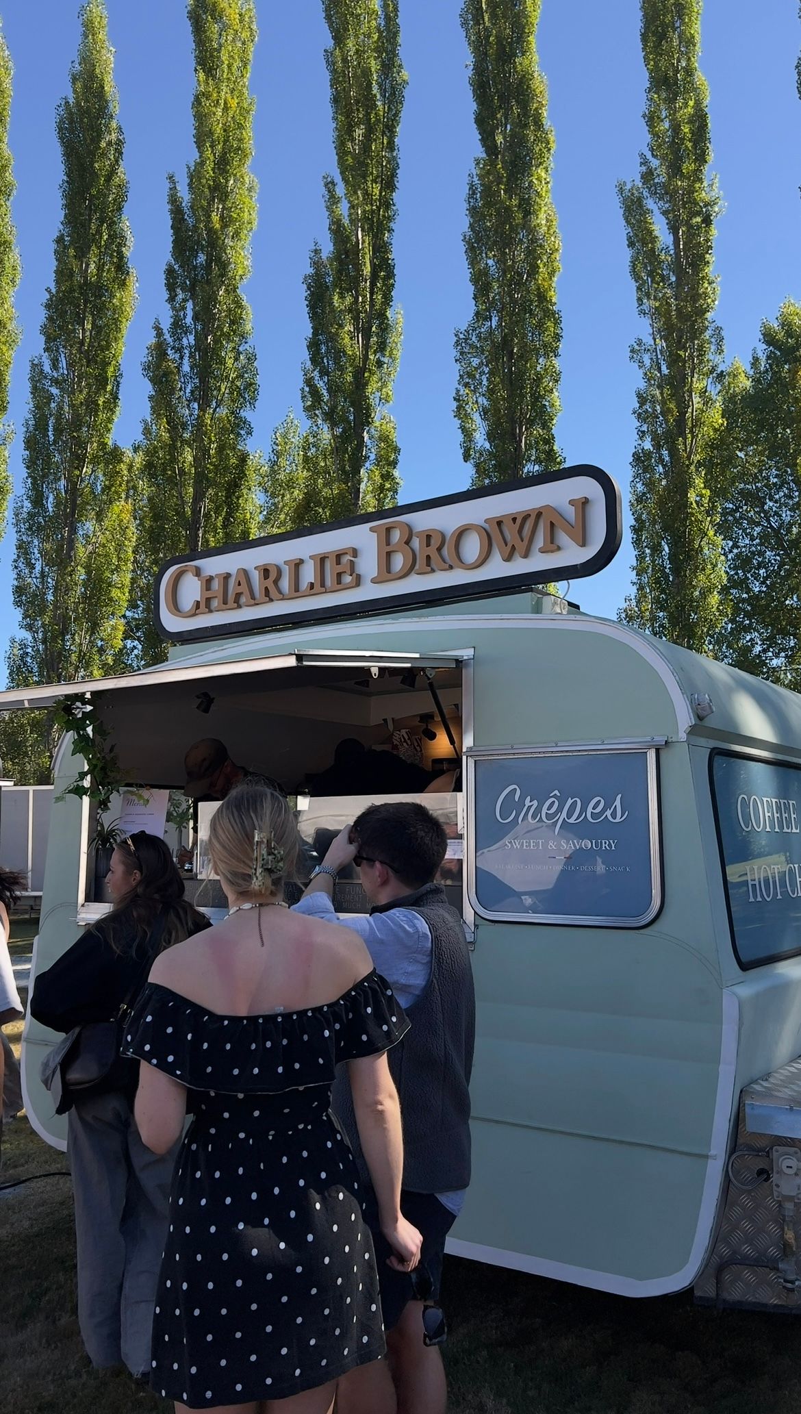 A line of customers wait at a white food truck named Charlie Brown's serving crepes under tall poplar trees.