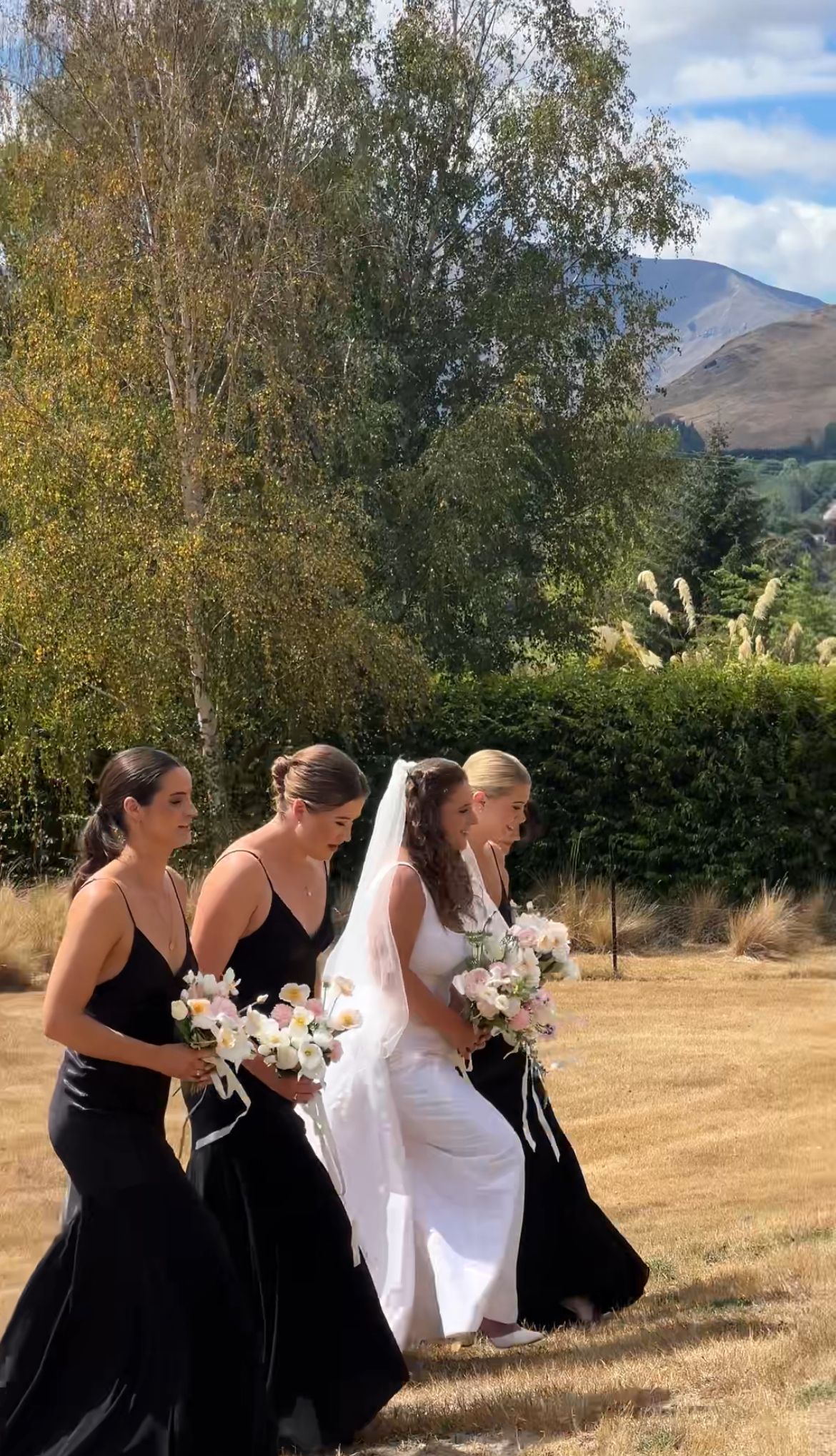 A wedding party walks across a grassy field with mountains and autumn trees in the background.