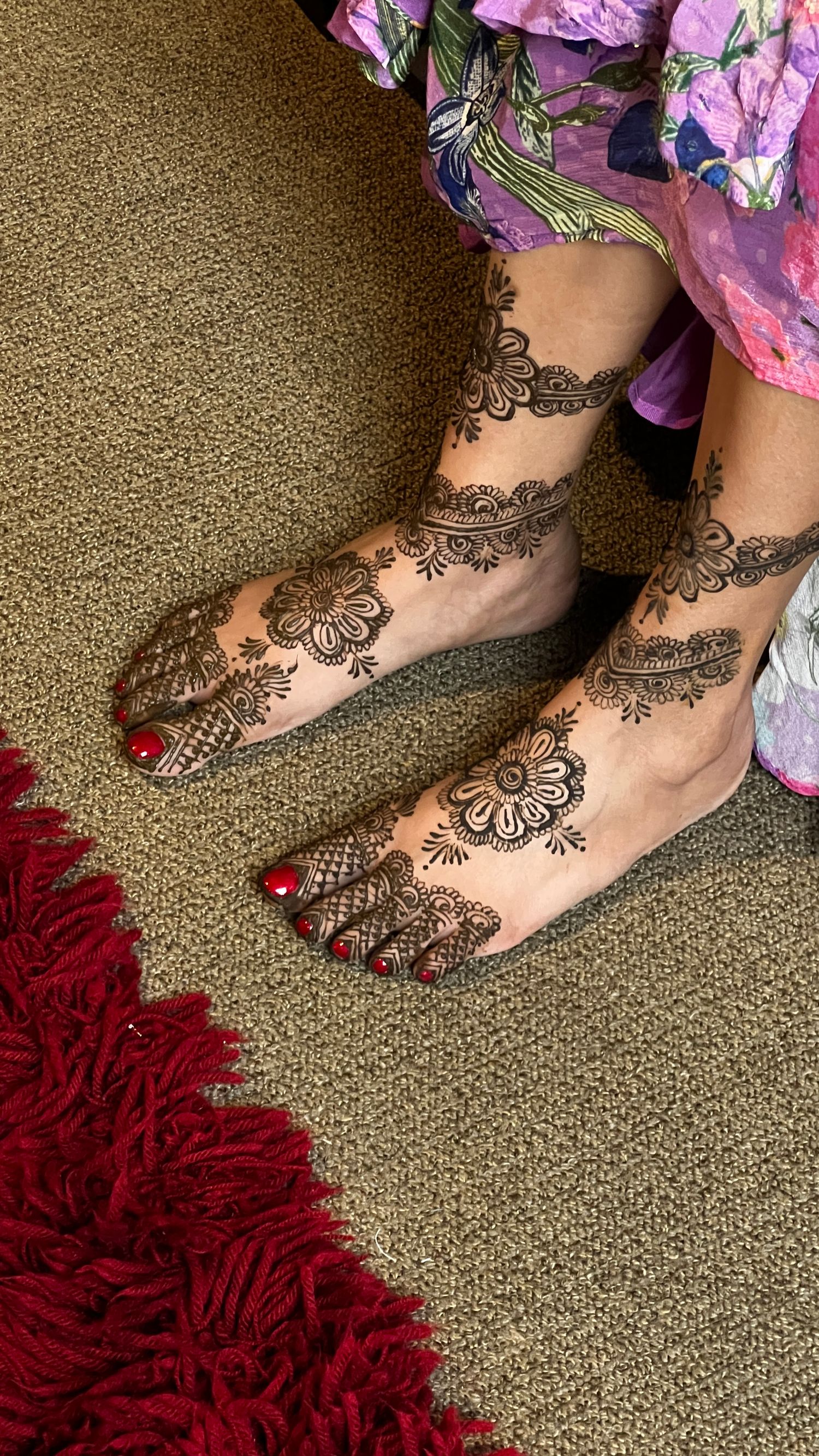 Detailed henna mehndi designs adorning feet with red nail polish on beige carpet.