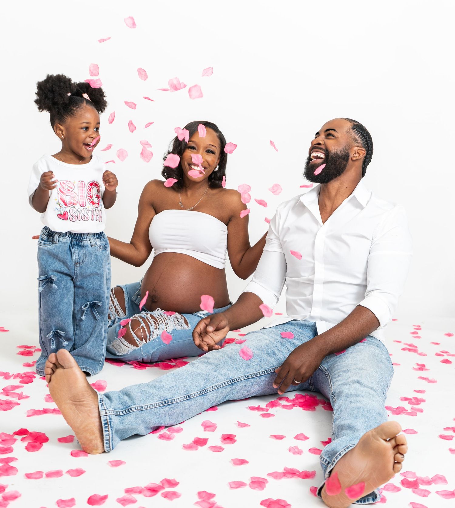 Playful maternity session in Vinings, Georgia — expecting black couple and daughter tossing pink rose petals in a bright studio.
