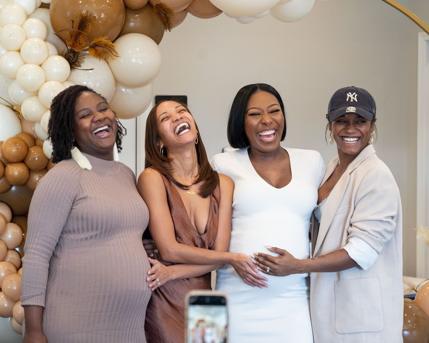 Baby-shower celebration in Roswell, Georgia — group of women laughing and embracing the expectant mother in front of a neutral balloon arch.