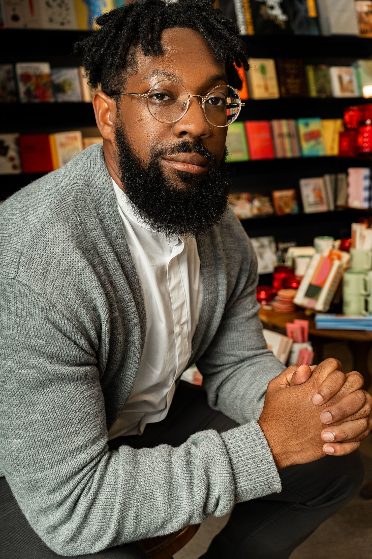 Confident portrait of a Black man with beard and glasses, captured indoors in a warmly lit bookstore setting — showcasing personality, style