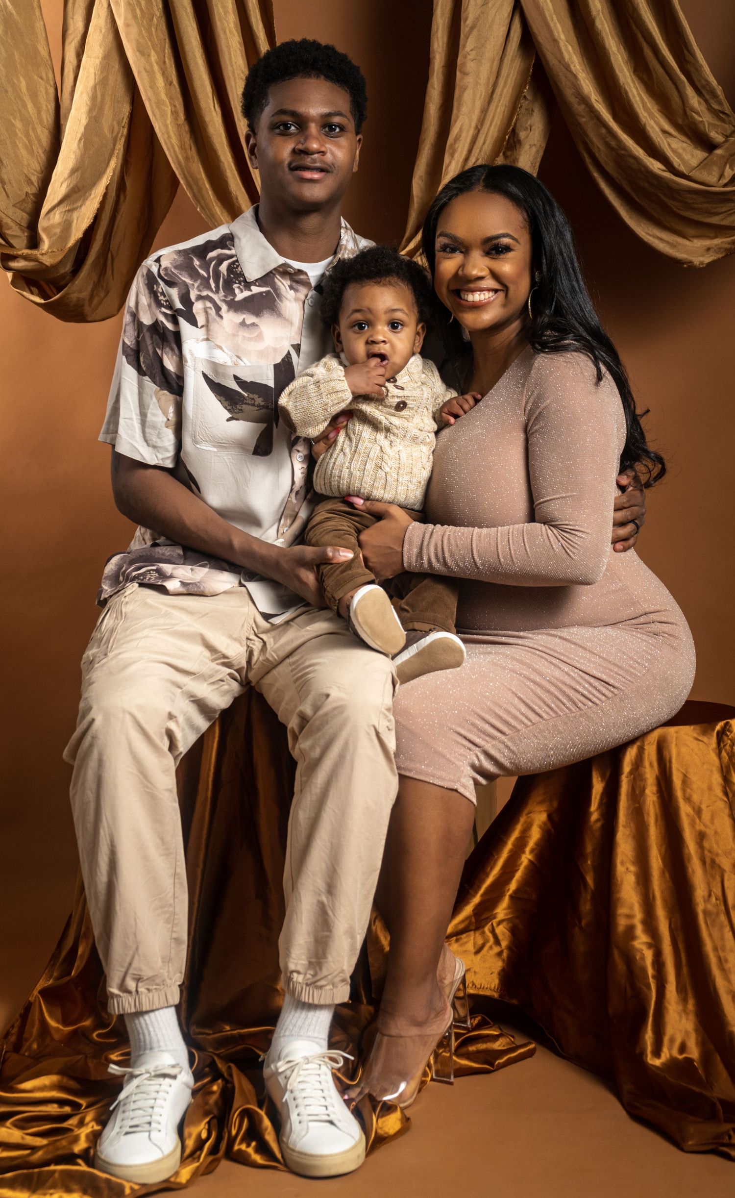 Warm family portrait featuring a young Black family of three posing in a beautifully styled studio with a brown-themed backdrop.