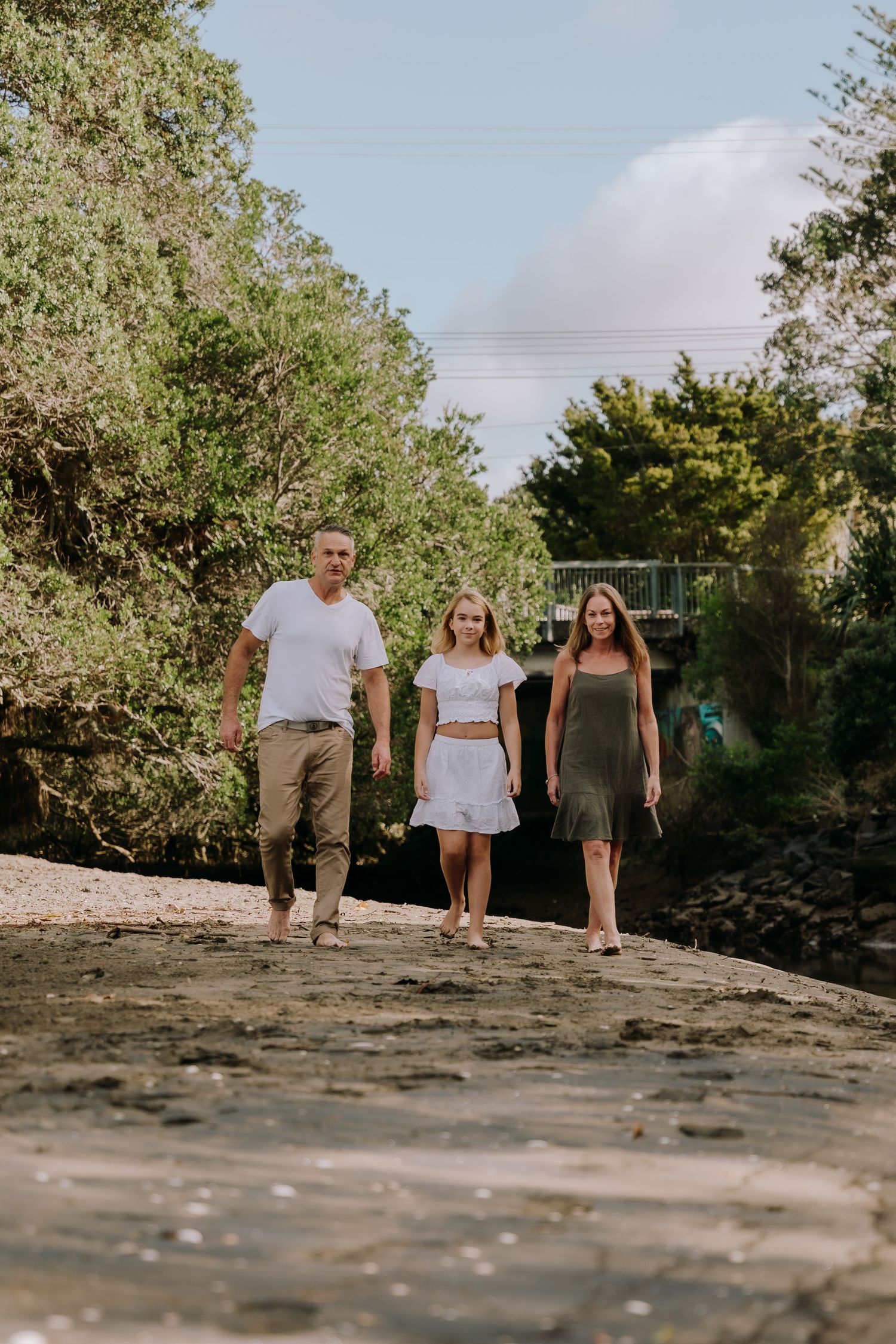Three people walking together on a rocky beach path surrounded by lush green trees on a sunny day.