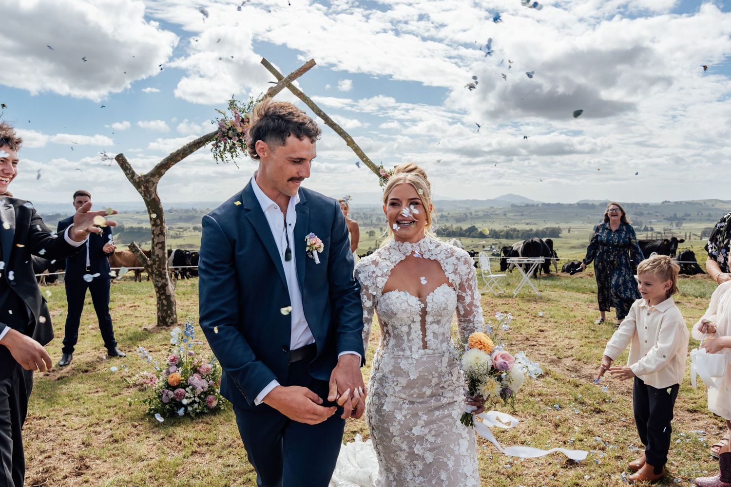 Newlyweds walk through confetti shower after outdoor wedding ceremony at rustic wooden arch with rolling hills backdrop.
