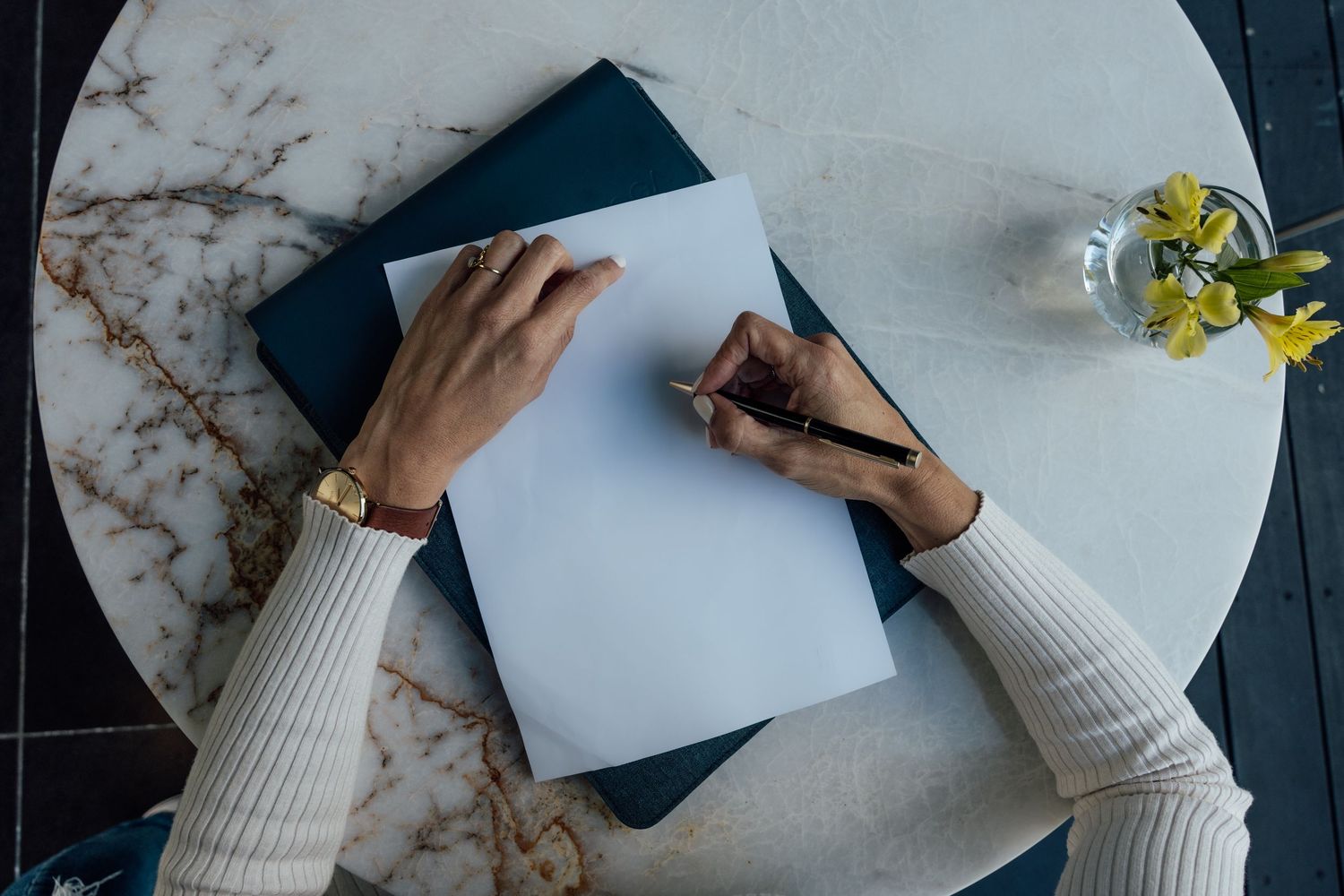 Woman writing on blank white paper in a dark blue notebook on a marble table with yellow flowers.