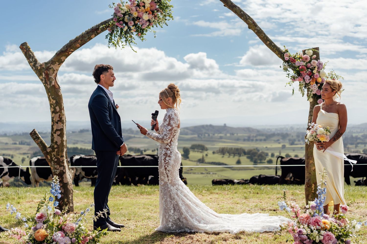 Outdoor farm wedding ceremony under floral arch with bride, groom, and officiant, rolling green hills and cattle in background.