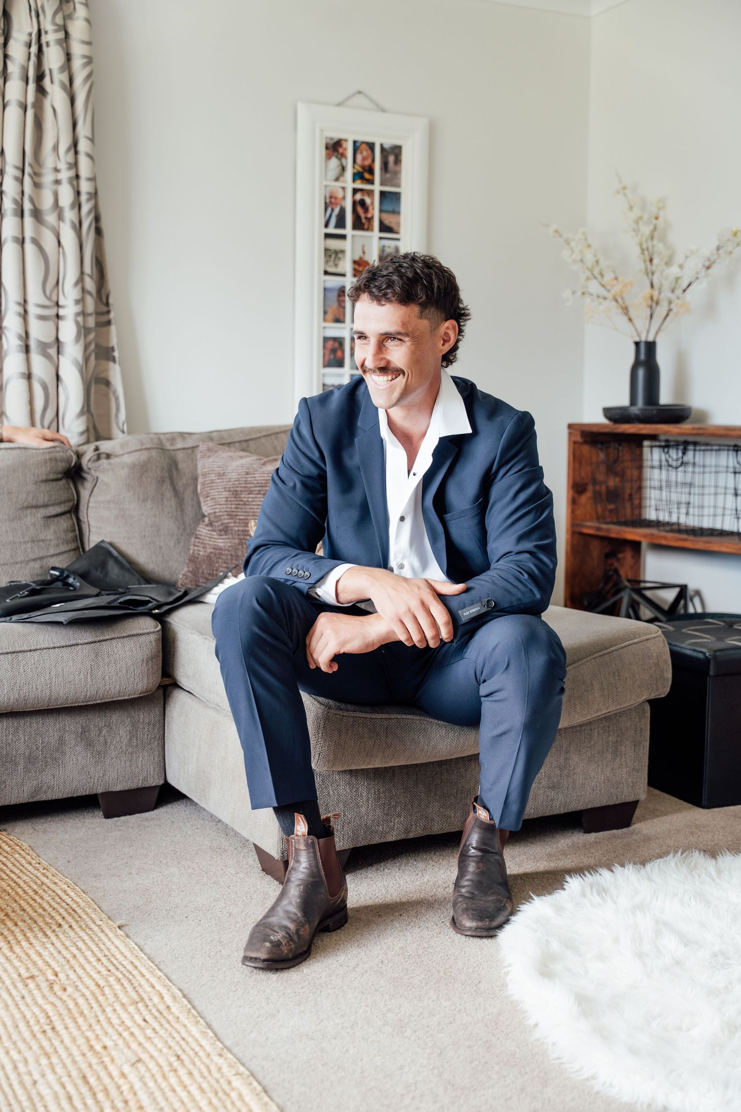 Smiling man in navy blue suit and brown boots sitting relaxed on a gray sofa in a stylish living room.