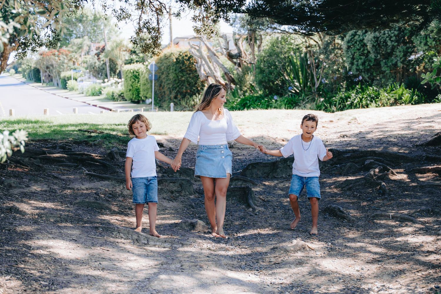 A mother holds hands with her two young sons in matching outfits while walking outdoors in a sunny park.