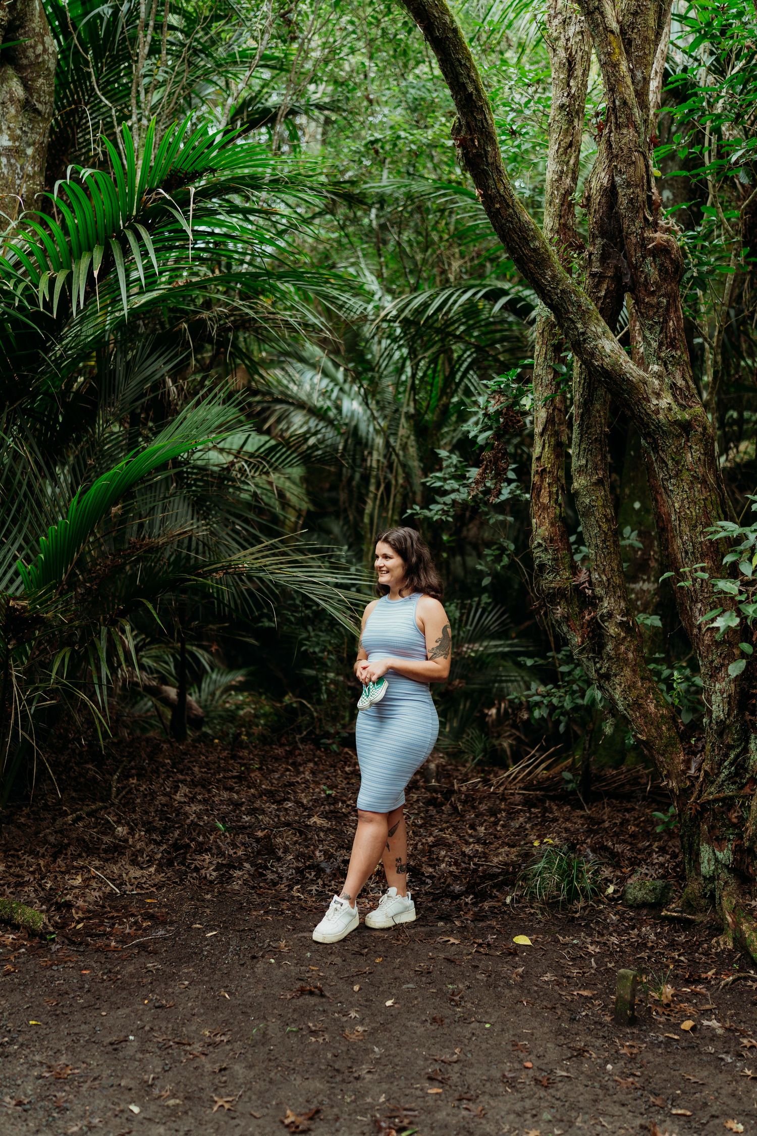 Woman in gray dress standing on forest path surrounded by lush tropical trees and palm fronds.