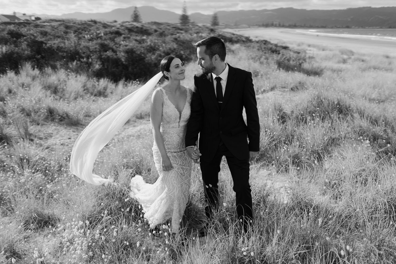 A black and white wedding photo of a couple standing in a field with bride's veil flowing in the wind.