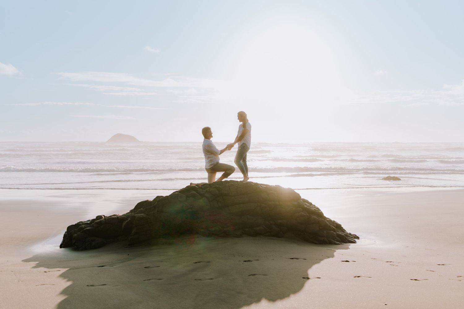 A romantic proposal scene silhouetted on a large rock formation at the beach during sunset.