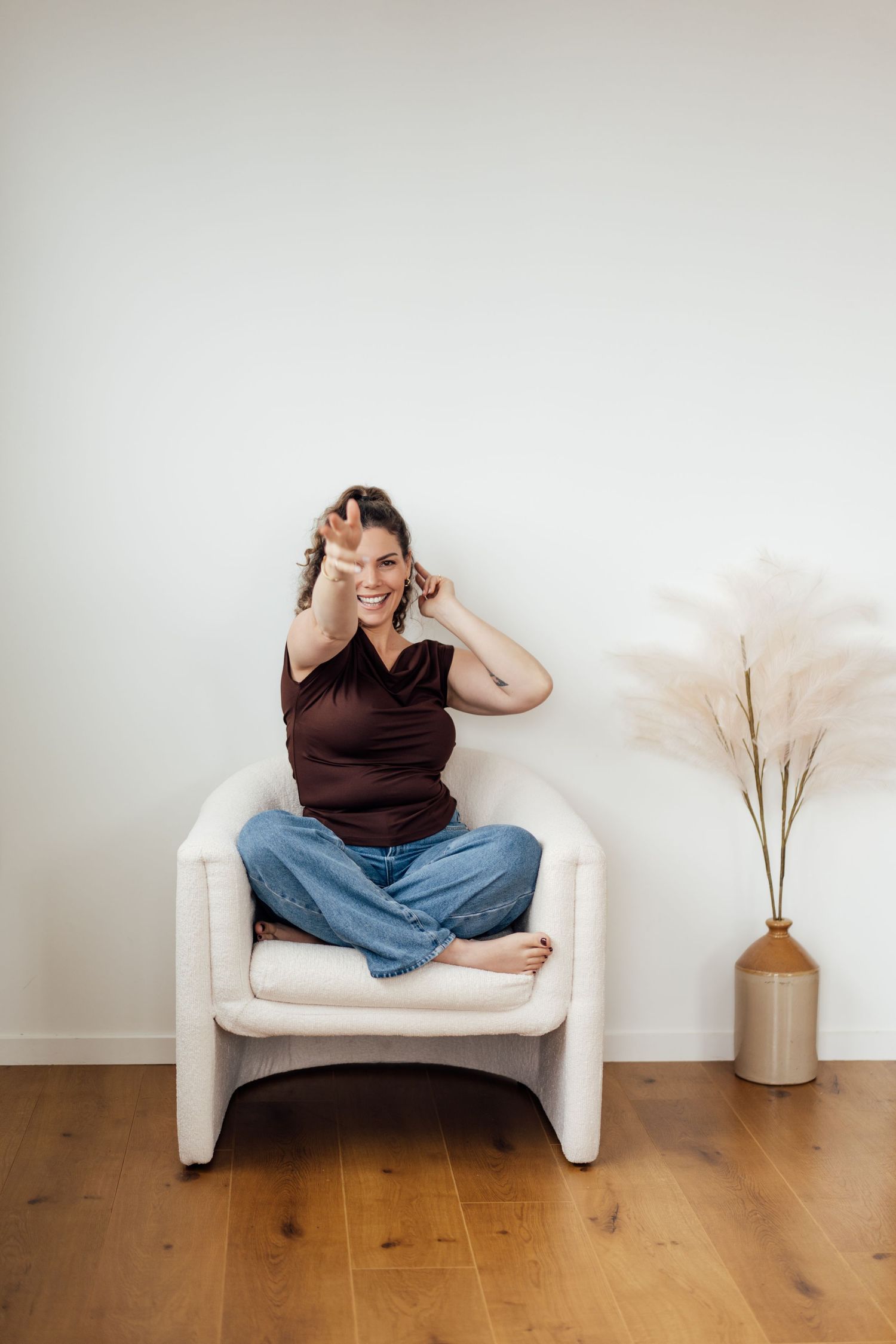 Woman sitting cross-legged in white chair, making peace sign, wearing brown top and jeans, smiling playfully.