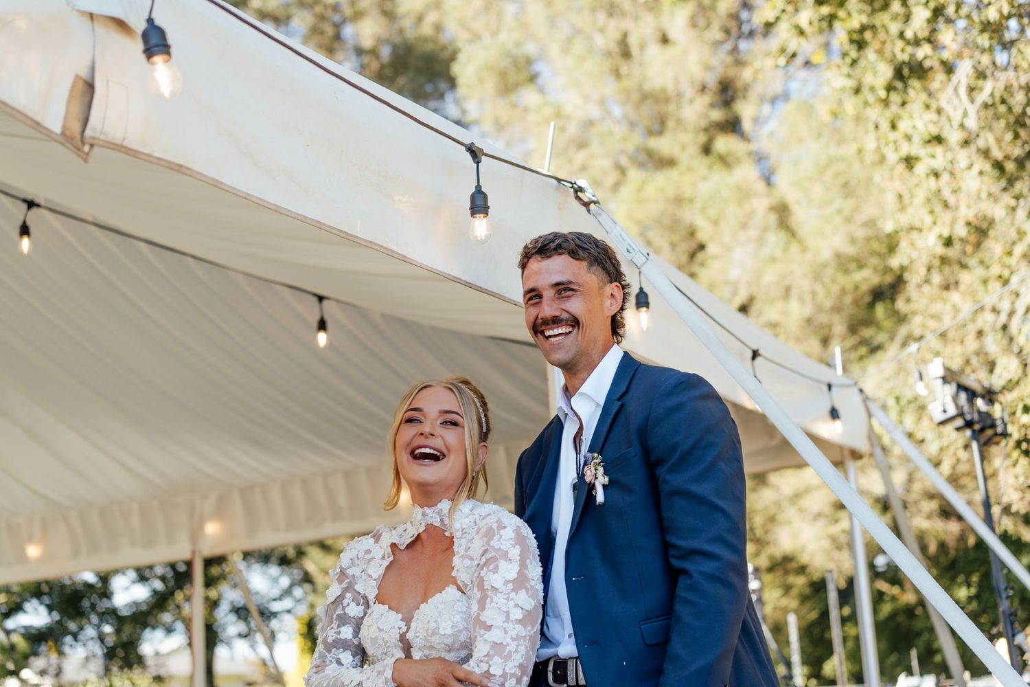 Happy bride and groom laughing outdoors under a white tent with string lights at their wedding reception.