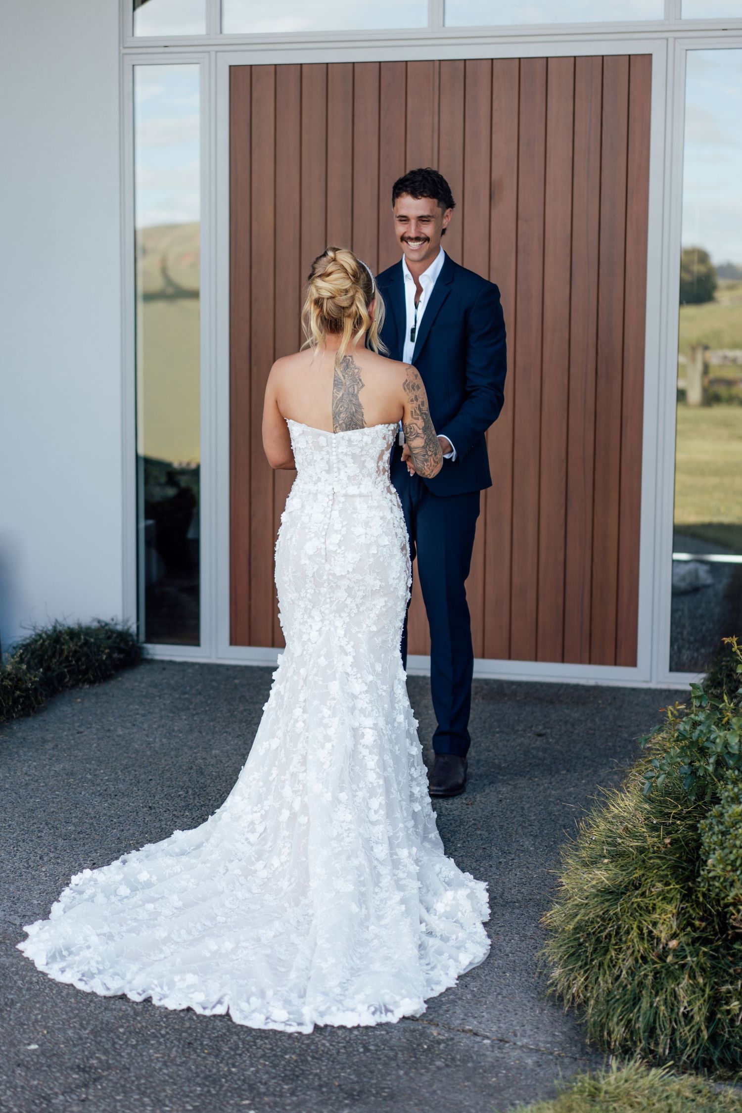 Bride in floral mermaid gown with back tattoo faces smiling groom in navy suit during outdoor first look.