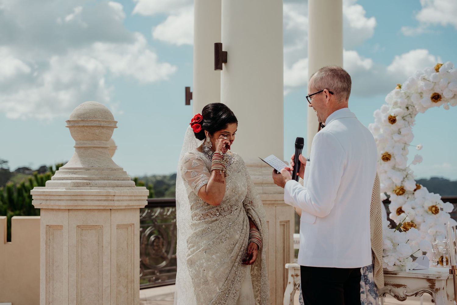 Bride in lace dress wipes tears during outdoor wedding ceremony at elegant venue with white floral arch.