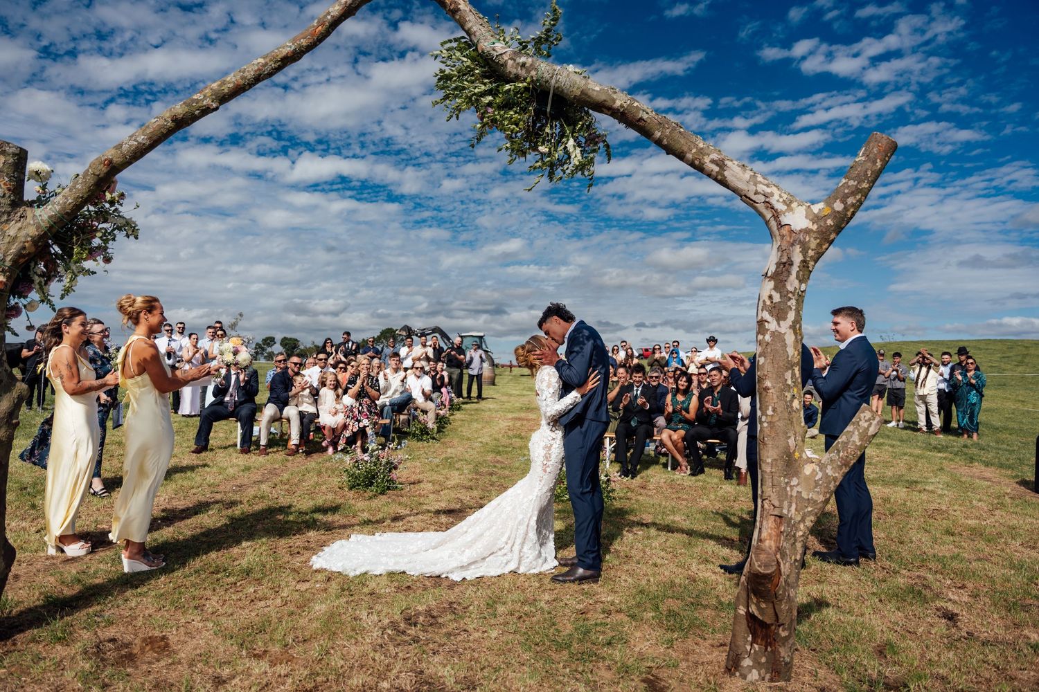 Bride and groom share first kiss at outdoor wedding ceremony under tree arch with guests gathered on green hillside.