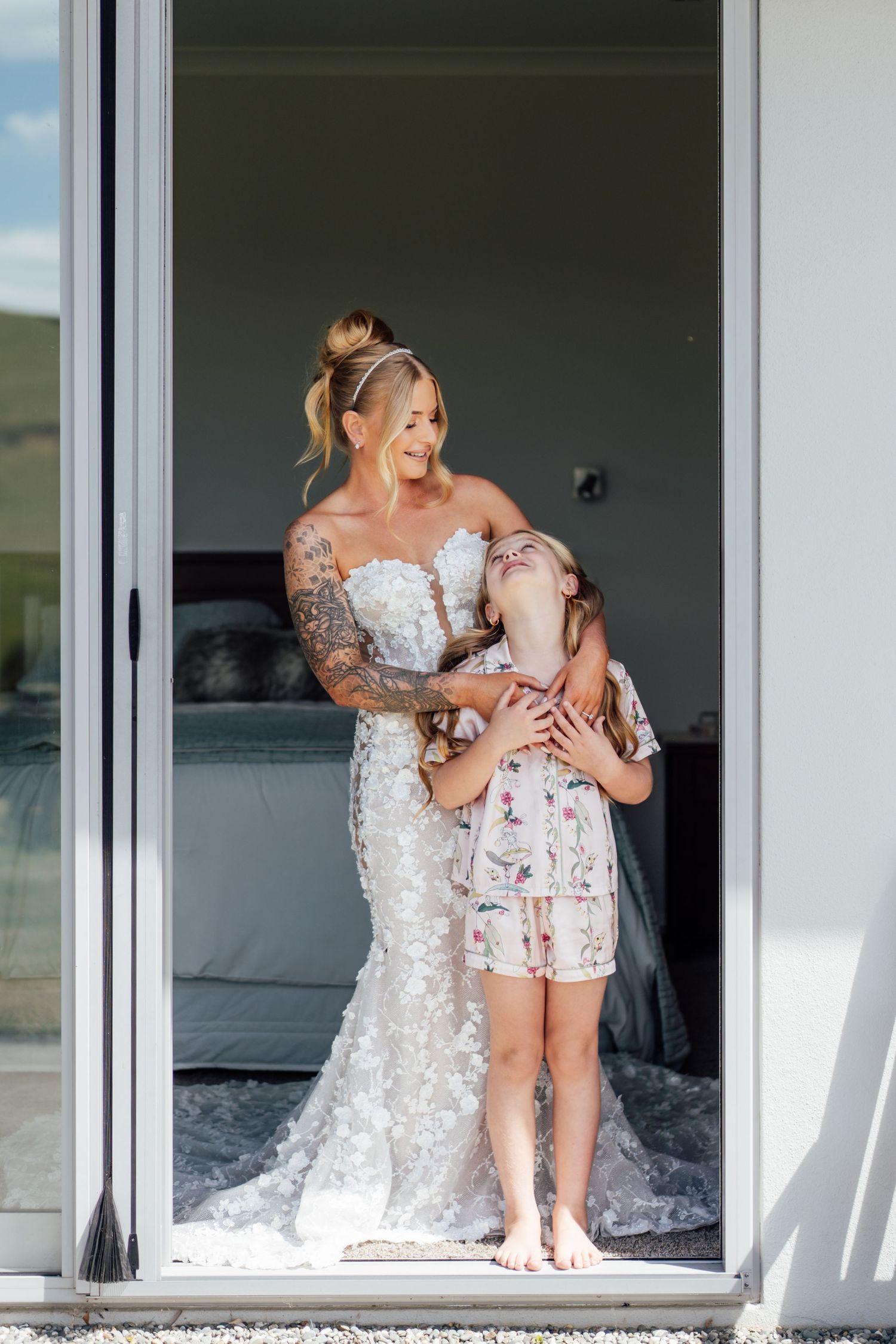 Bride in lace wedding gown hugging young girl in floral dress in sunlit doorway.