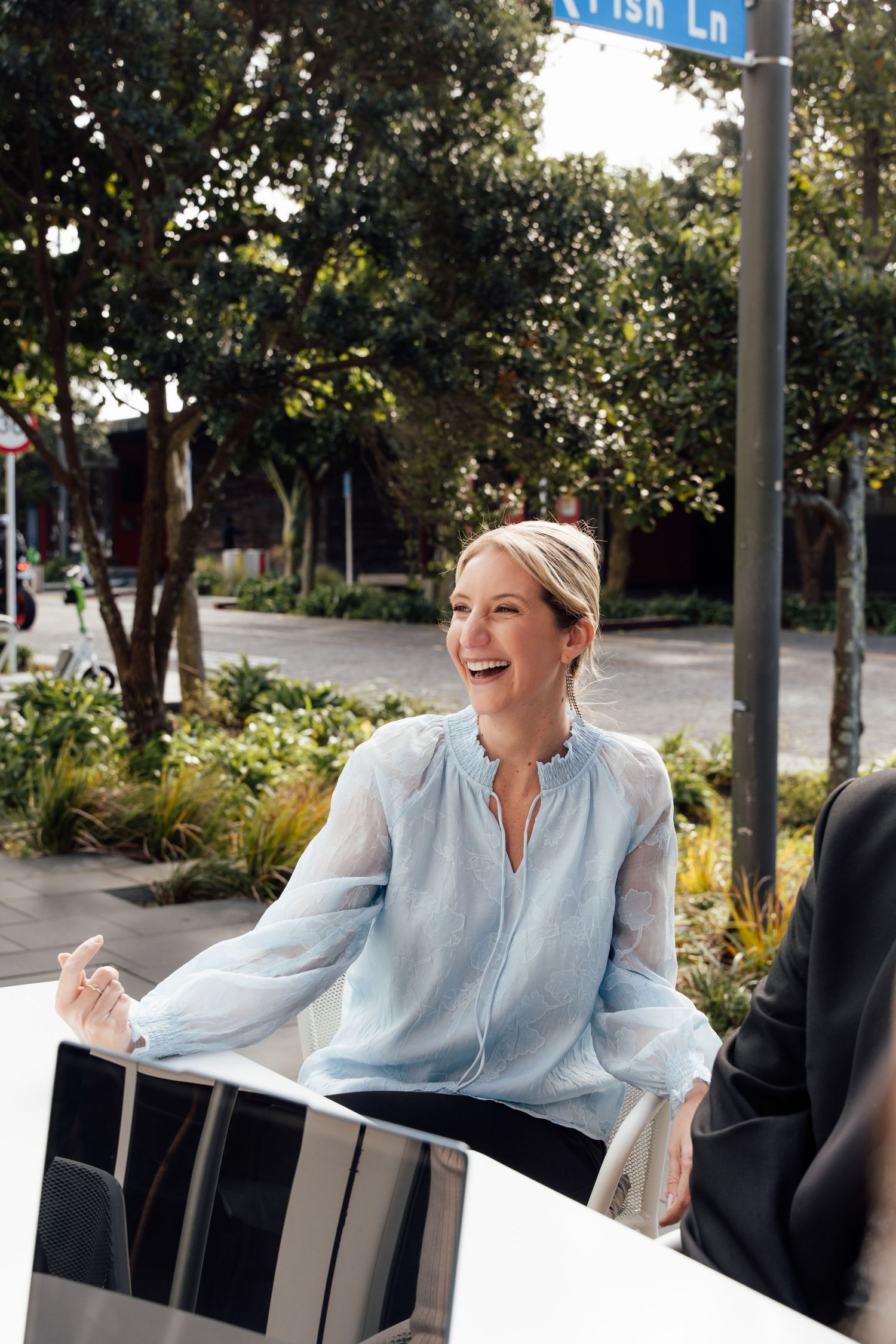 Woman in light blue blouse laughing outdoors at a cafe table on a sunny urban street.