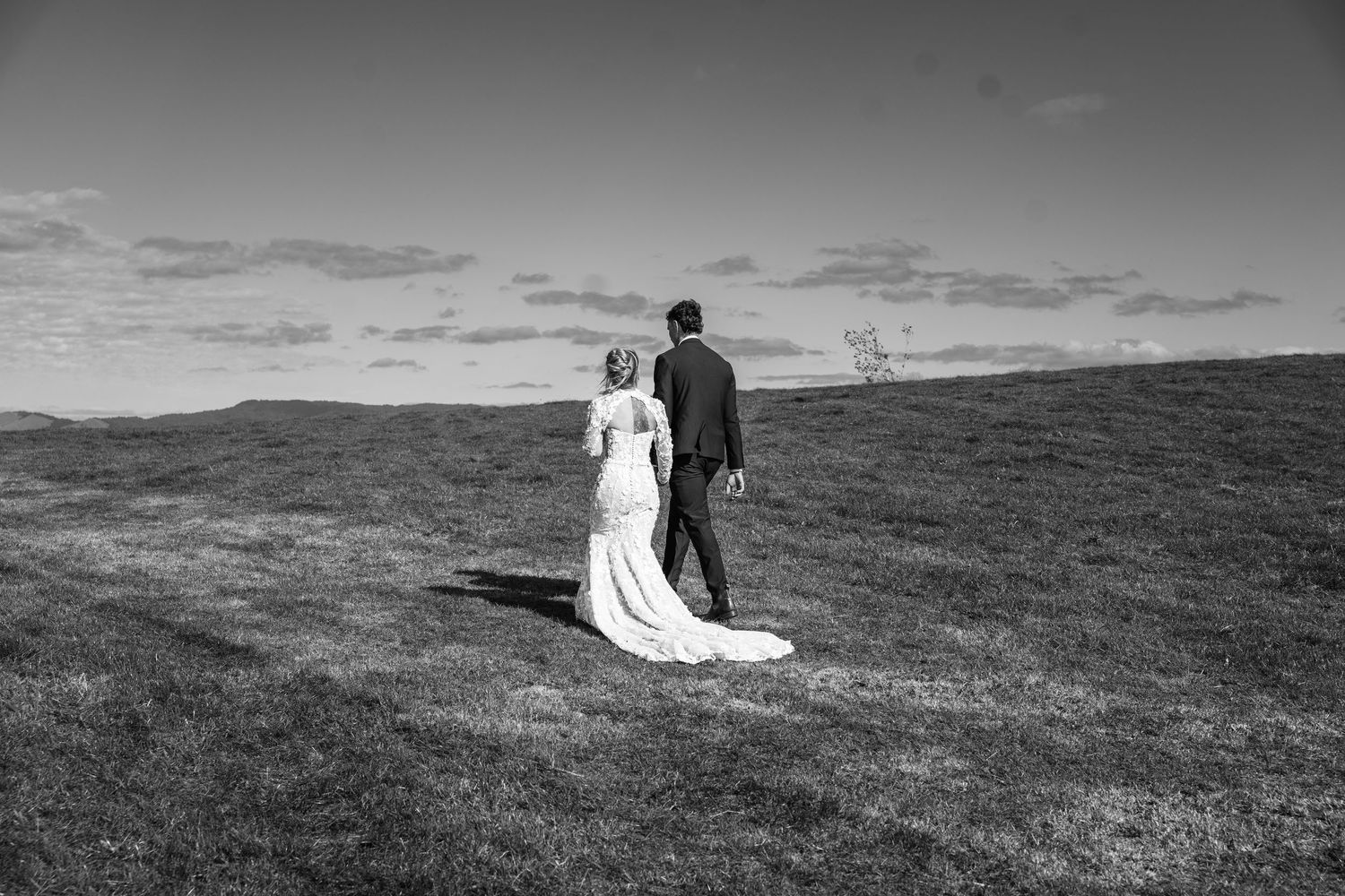 Black and white photo of newlywed couple walking hand in hand across a scenic grassy hillside.