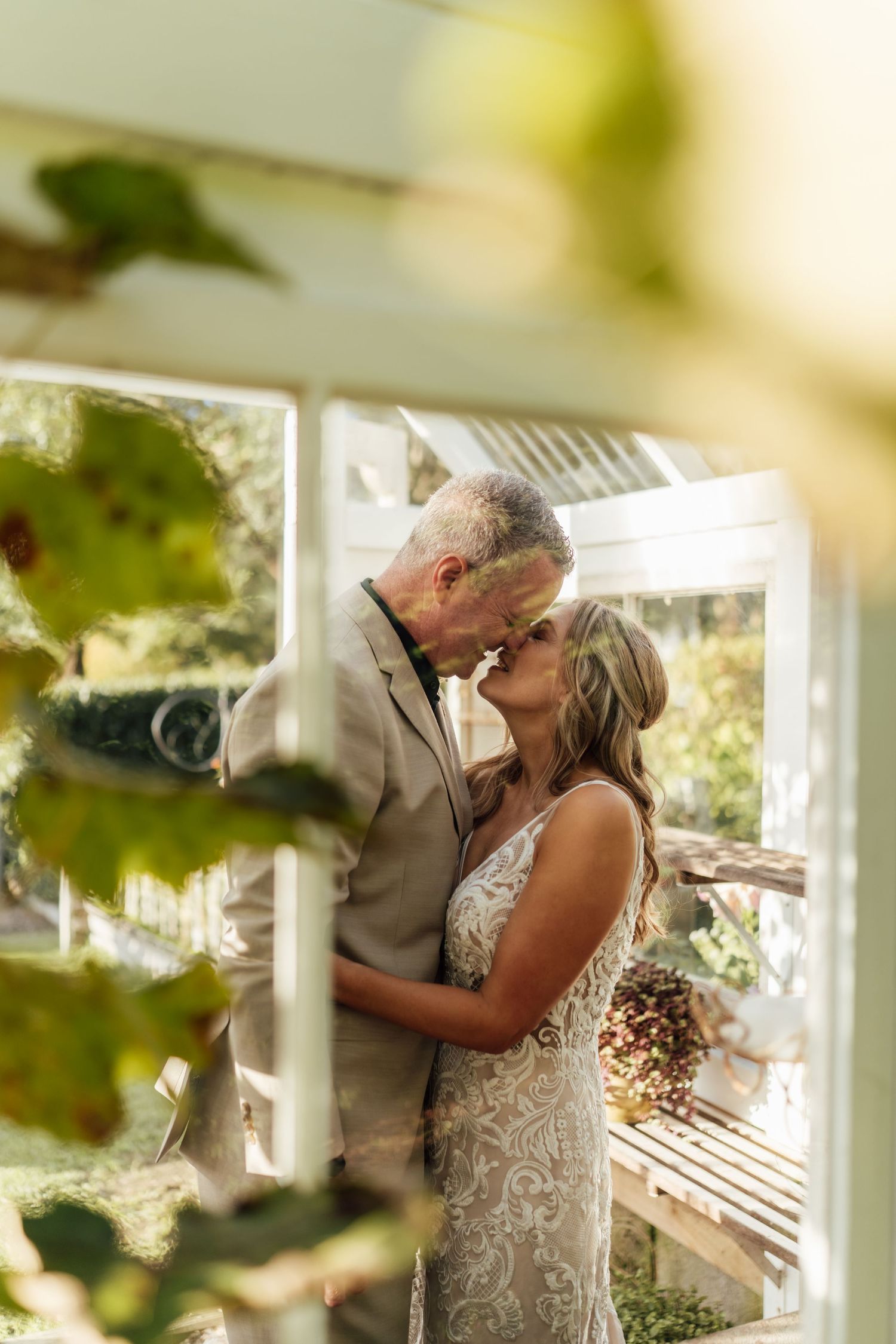 Bride and groom share a romantic kiss inside a sunlit greenhouse, surrounded by lush green foliage.