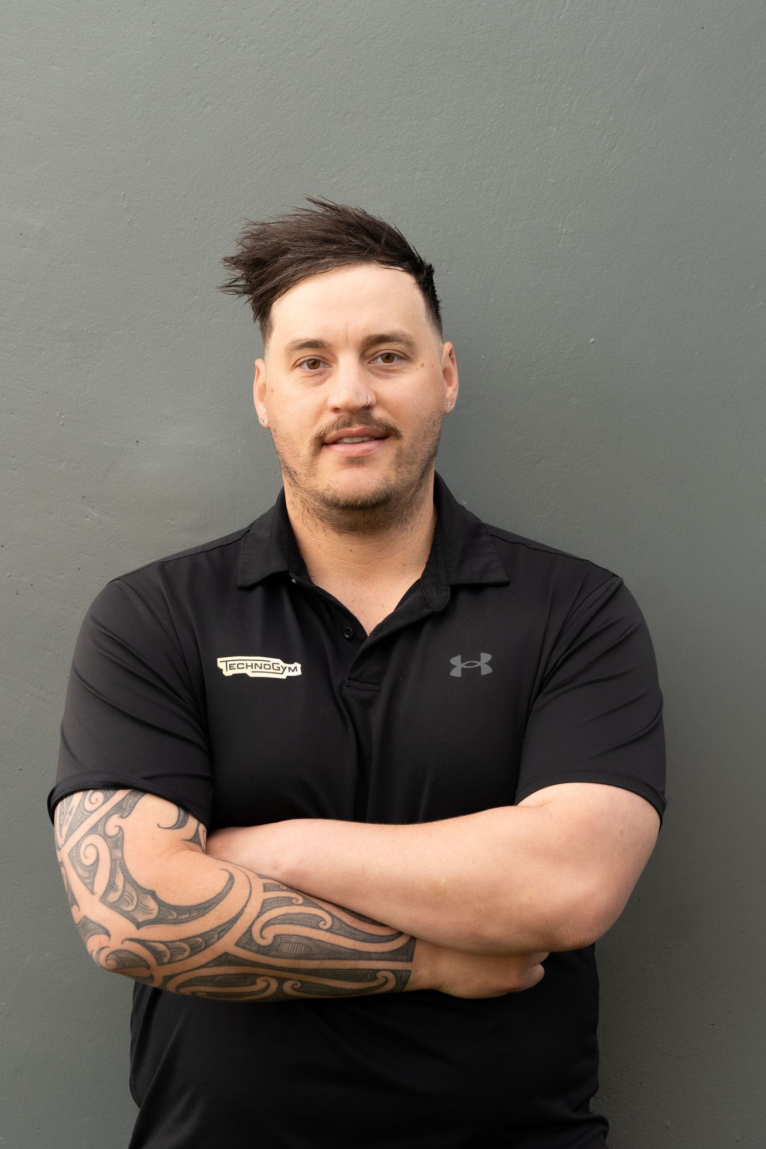 Man in black Under Armour polo with tribal tattoo, arms crossed, smiling against gray wall.