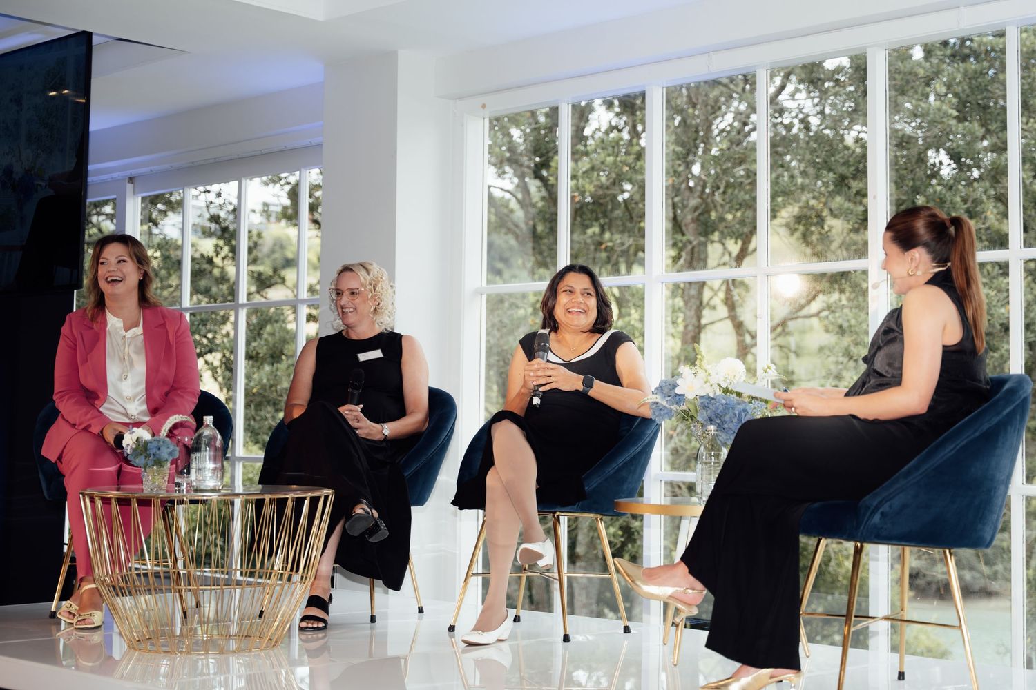 Four women seated in blue chairs during a panel discussion in a bright, modern room with large windows.