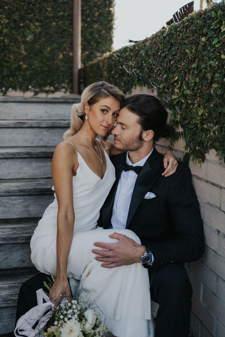 Elegant couple in wedding attire share an intimate moment on stone steps with greenery backdrop.
