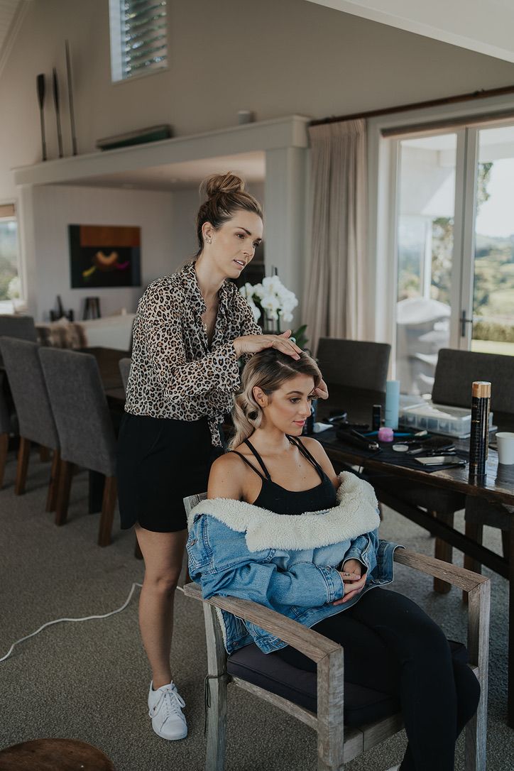 A hairstylist works on styling in a modern dining room with a window view and elegant grey chairs.