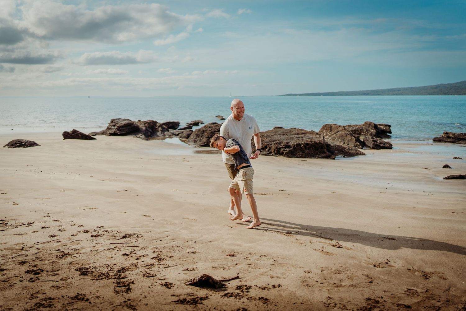 A person walks along a scenic sandy beach with rocky outcrops and blue ocean waters under a cloudy sky.