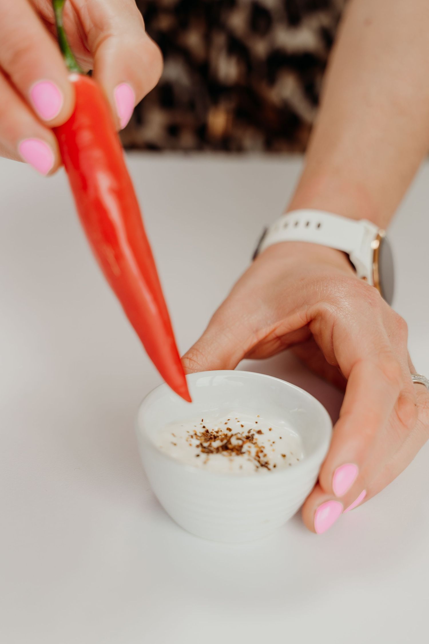 Woman with pink nails dipping a red chili pepper into a small white bowl of dip with black pepper.