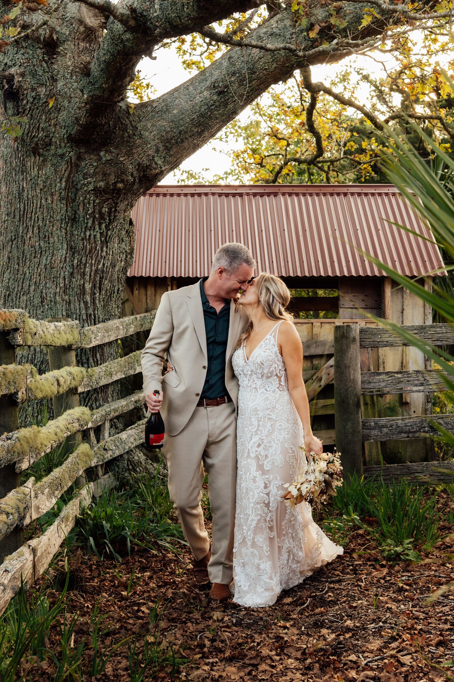 Bride and groom walk together on a rustic farm path, holding champagne and a bouquet under a large oak tree.