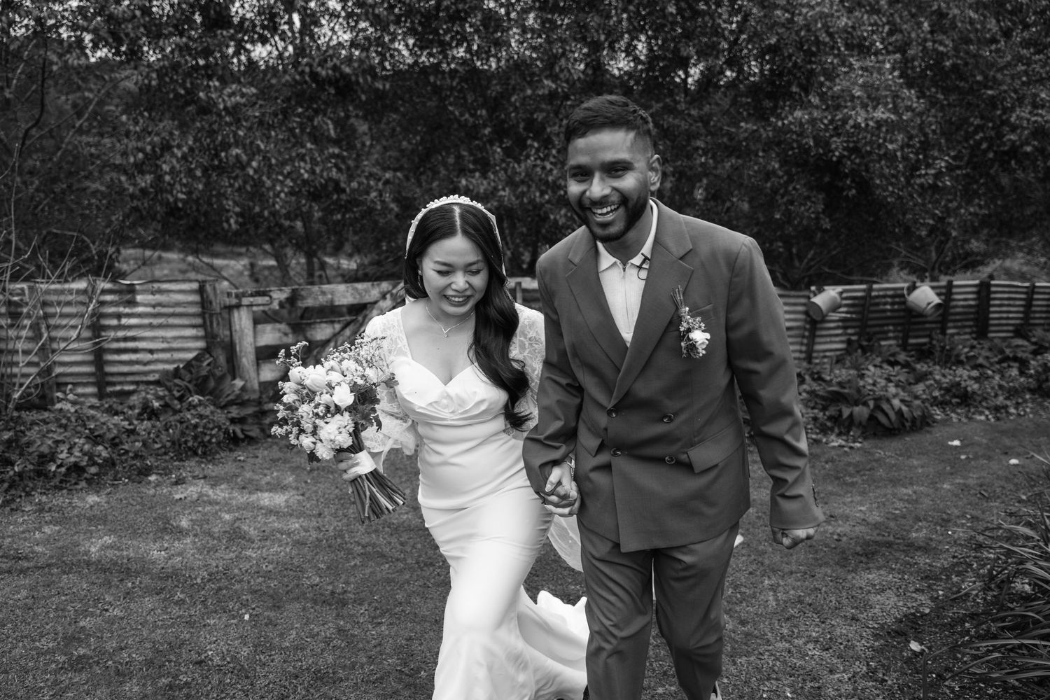 Black and white photo of a smiling interracial couple walking hand in hand outdoors on their wedding day.