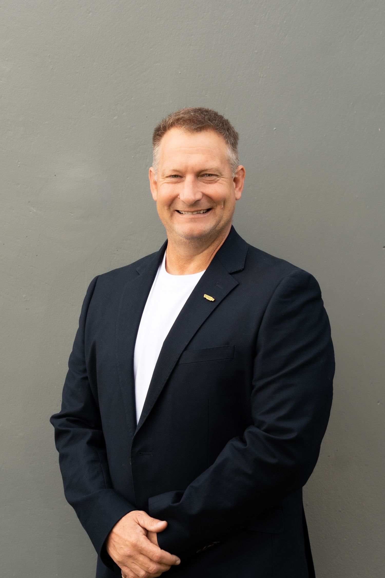 Smiling middle-aged man in dark blazer and white shirt poses confidently against a gray wall.