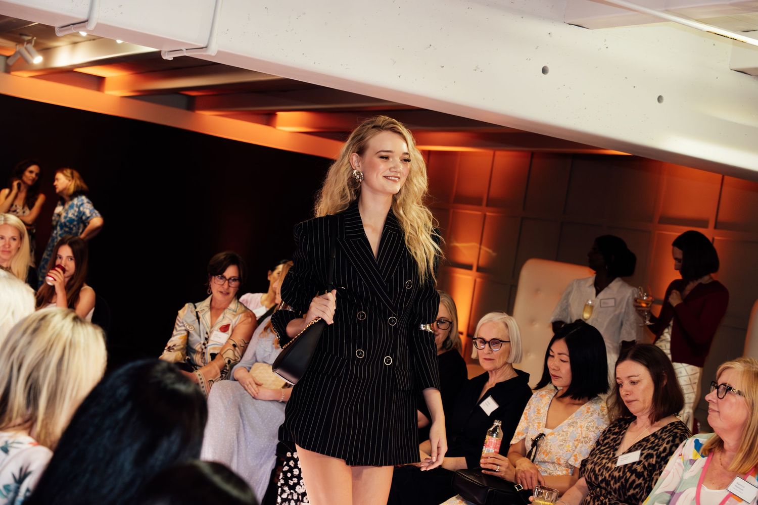 Model walks runway in black pinstripe blazer dress at fashion show with seated audience.