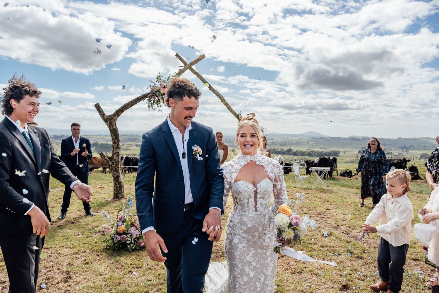 Joyful newlyweds walk down the aisle under a rustic wooden arch at an outdoor wedding ceremony with guests celebrating.