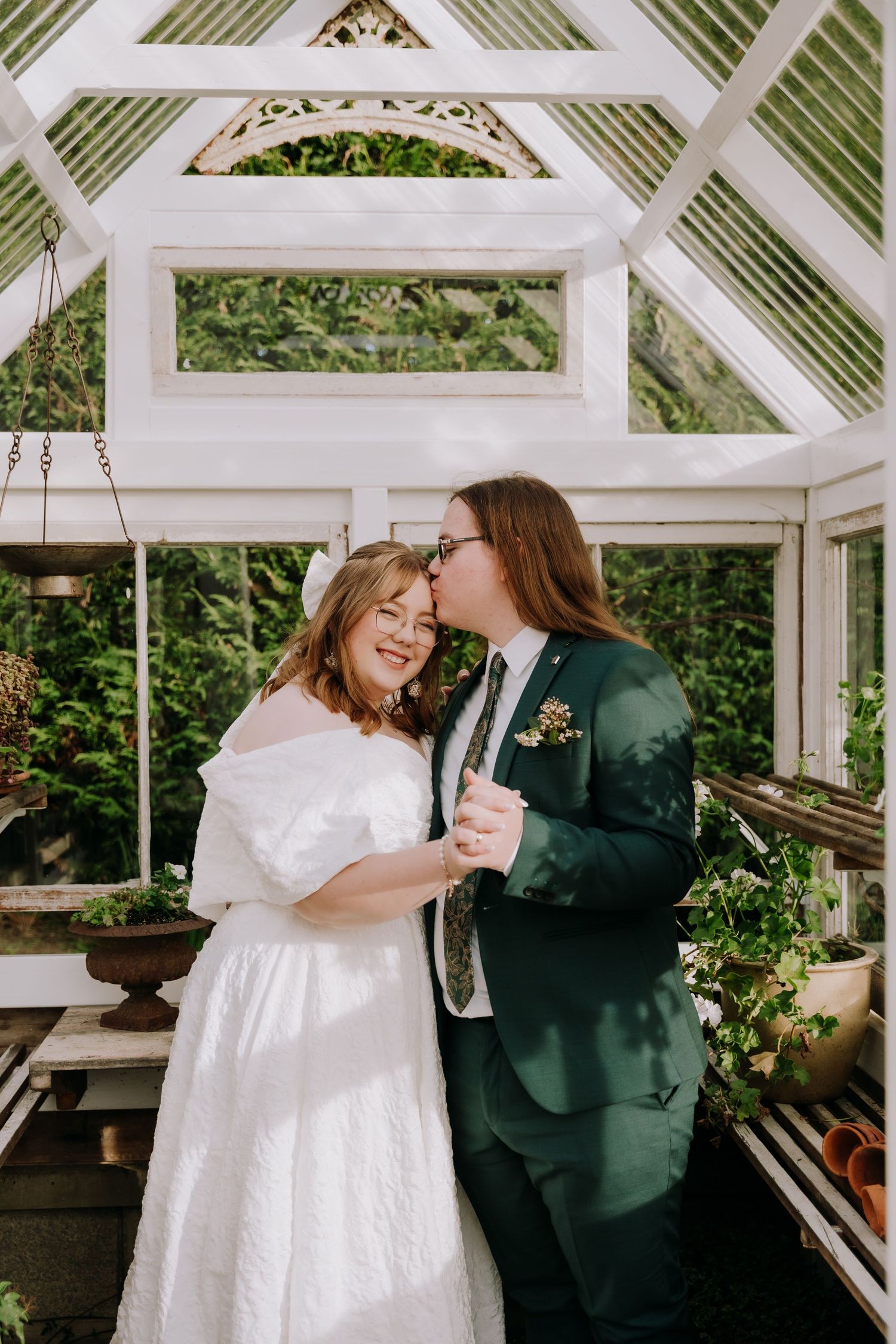 Newlyweds share a tender kiss in a sunlit greenhouse, bride in white gown, groom in green suit with boutonniere.