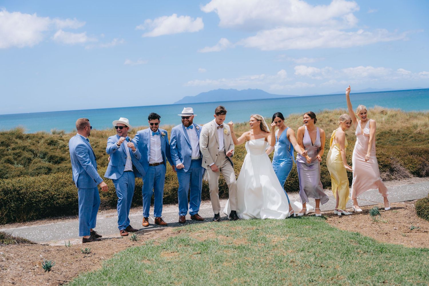 Wedding party poses outdoors near ocean, bride in white gown with groomsmen in blue suits and bridesmaids in pastel dresses.