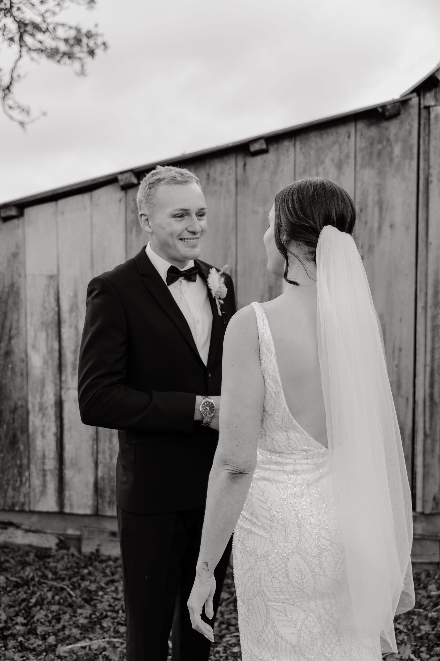 Black and white wedding photo showing a couple in formal attire sharing a first look moment by a rustic barn wall.