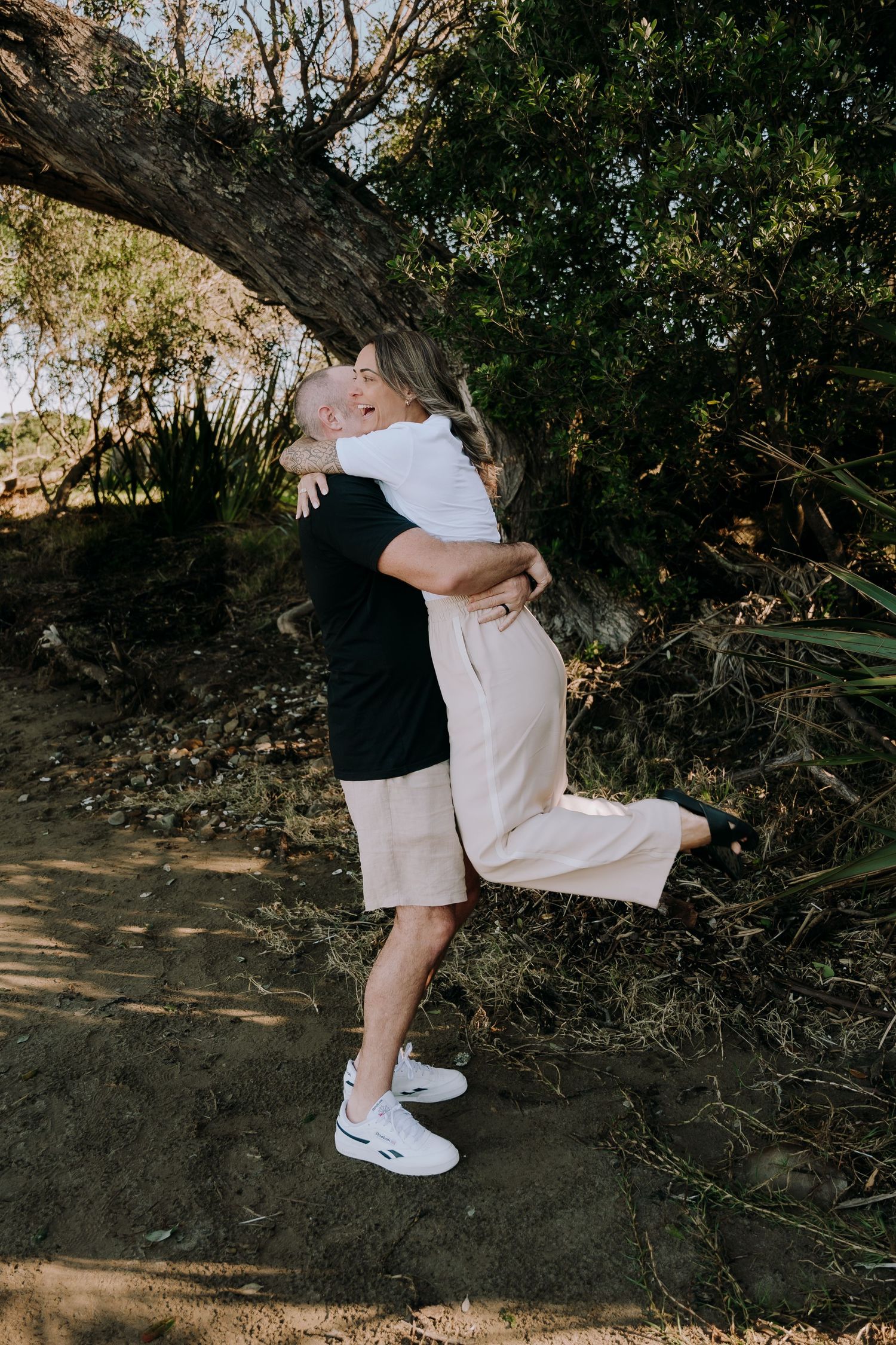 A couple in casual attire sharing an intimate embrace outdoors against a wooded backdrop.