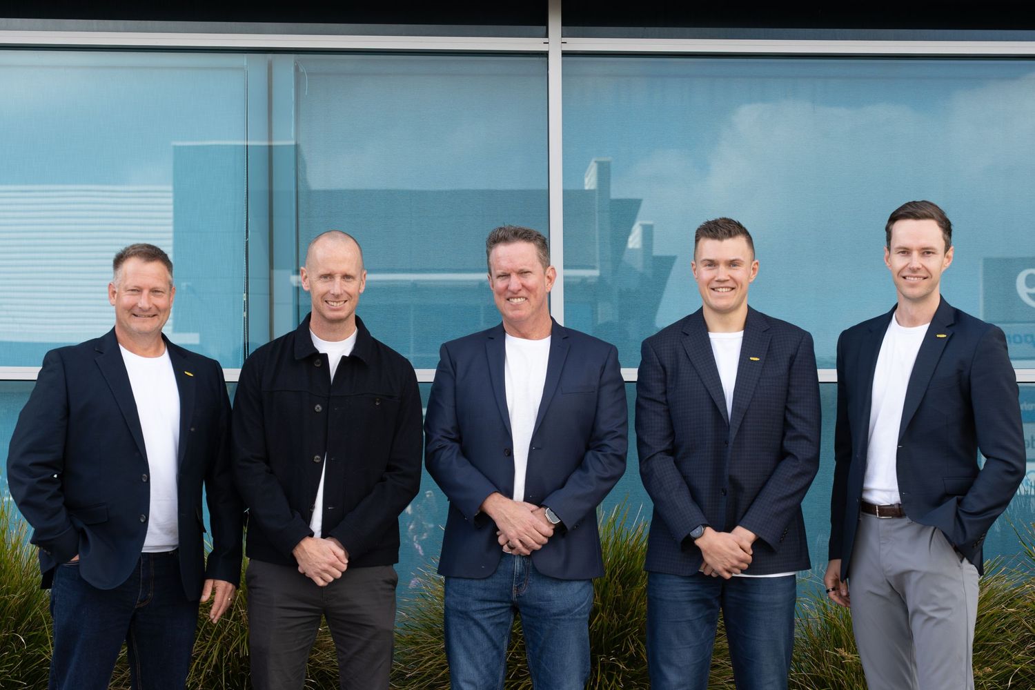 Five professional men in business casual attire posing together outdoors in front of a modern building.