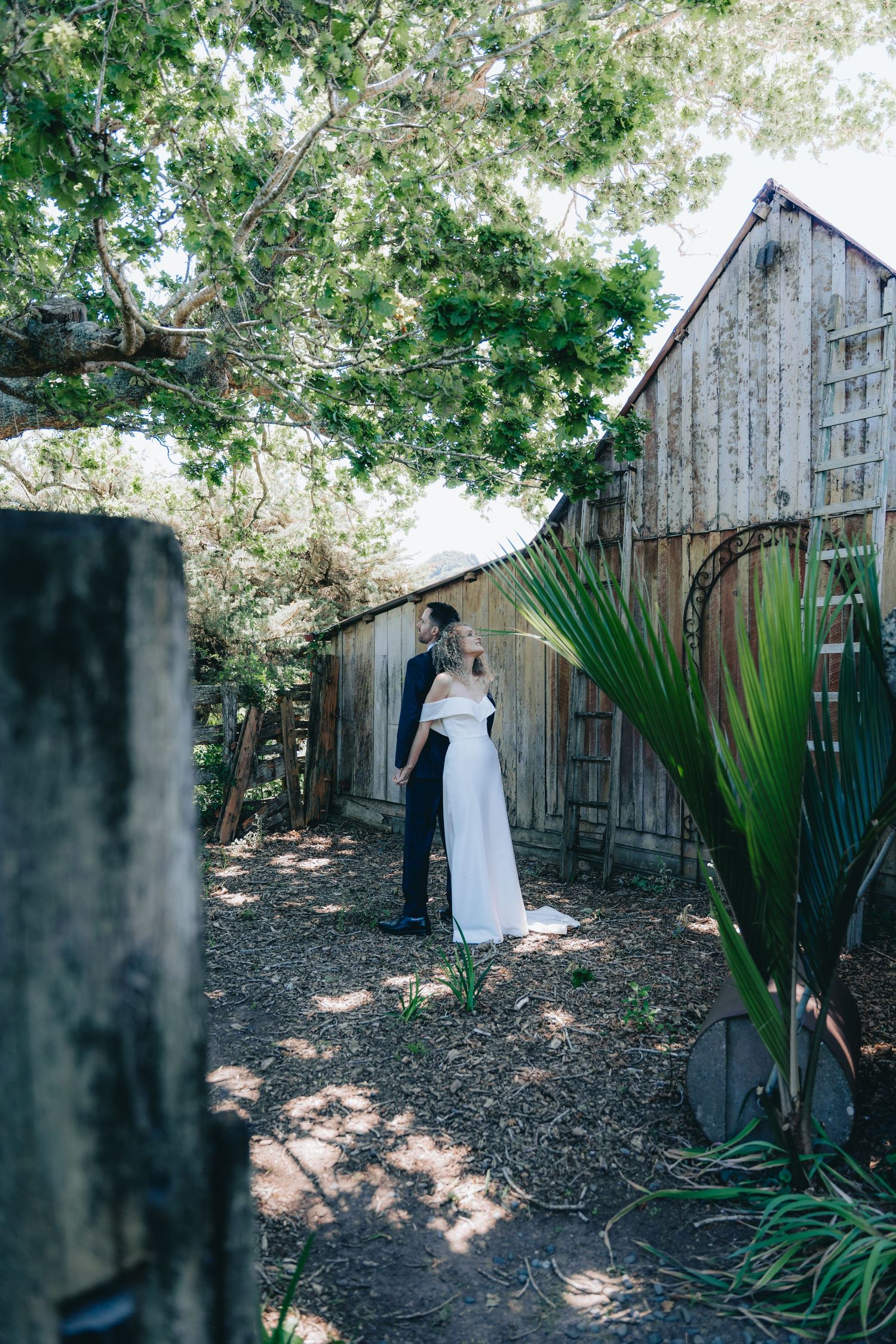 Bride and groom share a romantic moment in a rustic garden setting with wooden barn and tropical plants.